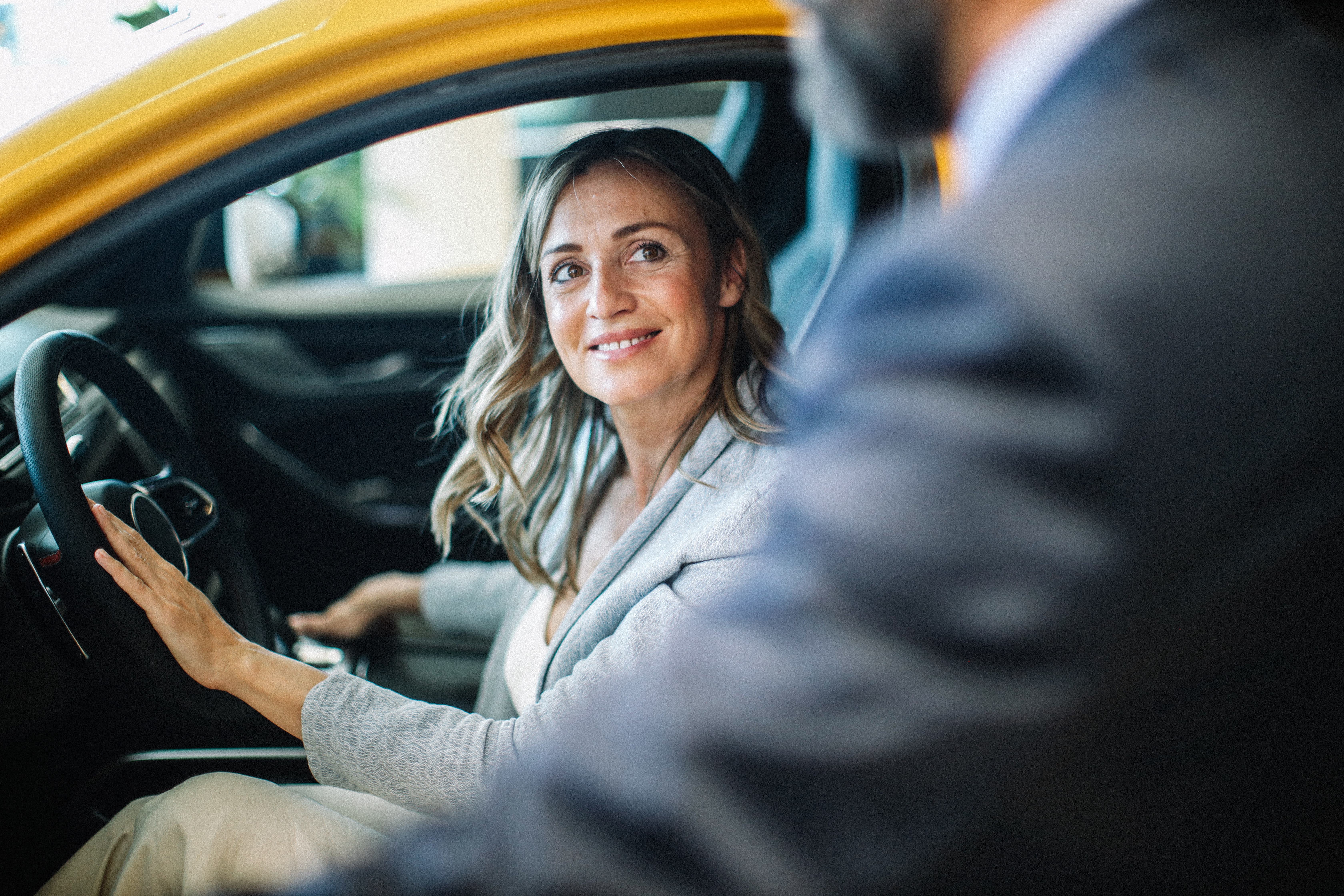 Ältere Frau sitzt in einem gelben Auto auf dem Fahrersitz und schaut auf eine Person, die im Vordergrund steht